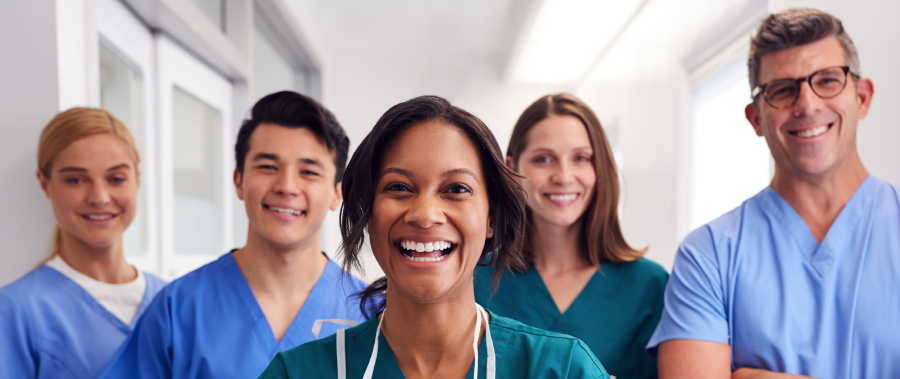 Five multi-cultural medical team members stand together in a hospital corridor. They are wearing scr