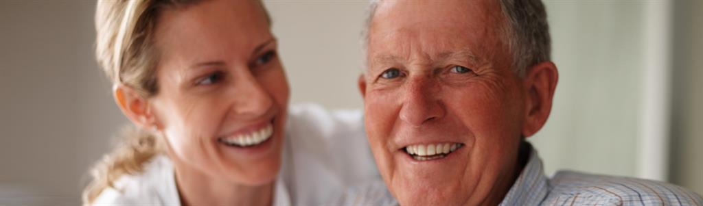 A smiling, elderly man sits in a wheelchair with a nurse hovering over his shoulder.