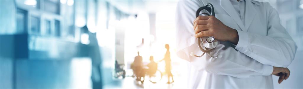 Physician in a white coat with stethoscope in hand. Patients in the background out of focus.